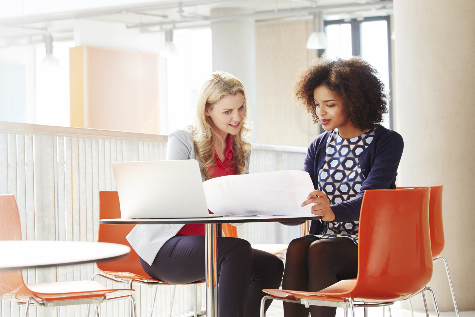 Two professionals sitting at a table looking at paper documents with a laptop on the table.