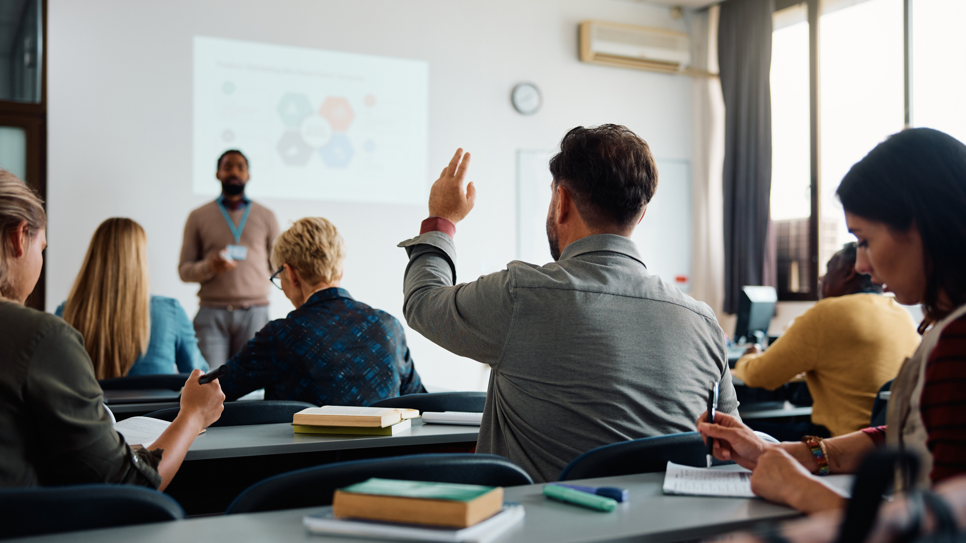 Diverse adult learners in classroom with man raising hand during instructor-led presentation with projected slides
