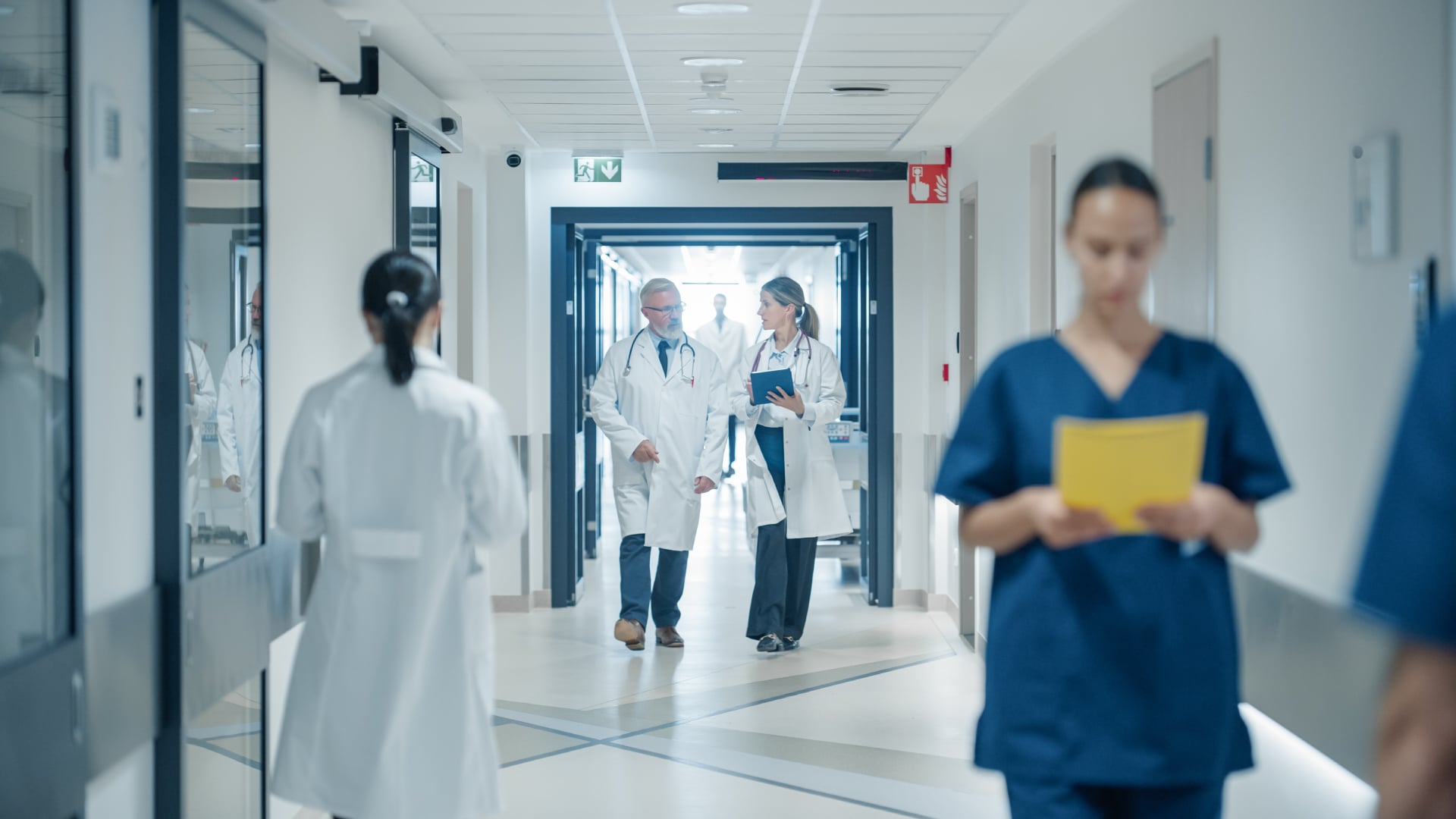 Doctors in white coats and nurses in scrubs walking through a busy modern hospital corridor