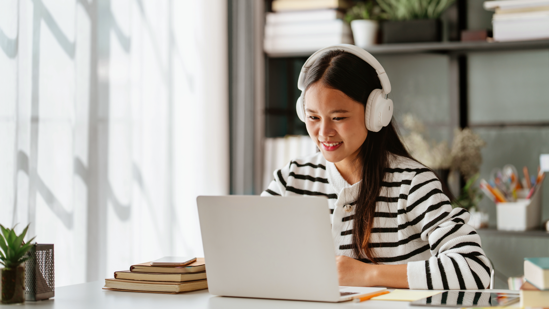 Young woman wearing white headphones smiling at laptop in bright home study with books and plants
