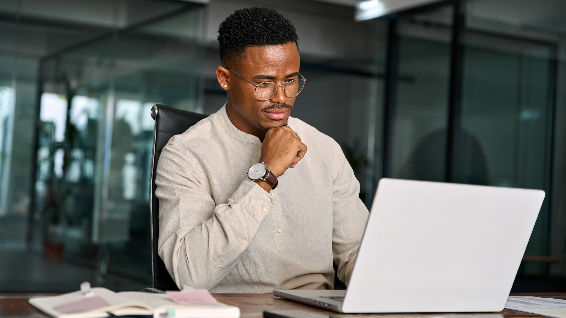 Young professional man with glasses reviewing laptop at office desk with hand on chin