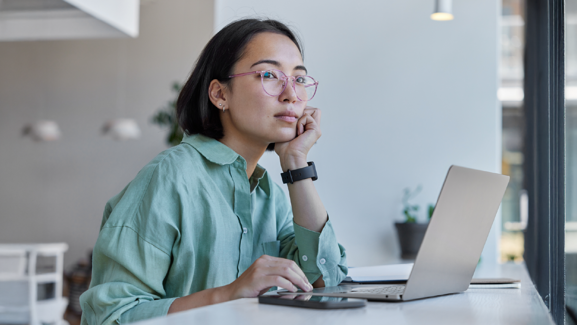 Young woman with pink glasses gazing away from laptop in modern office, appearing distracted or uncertain