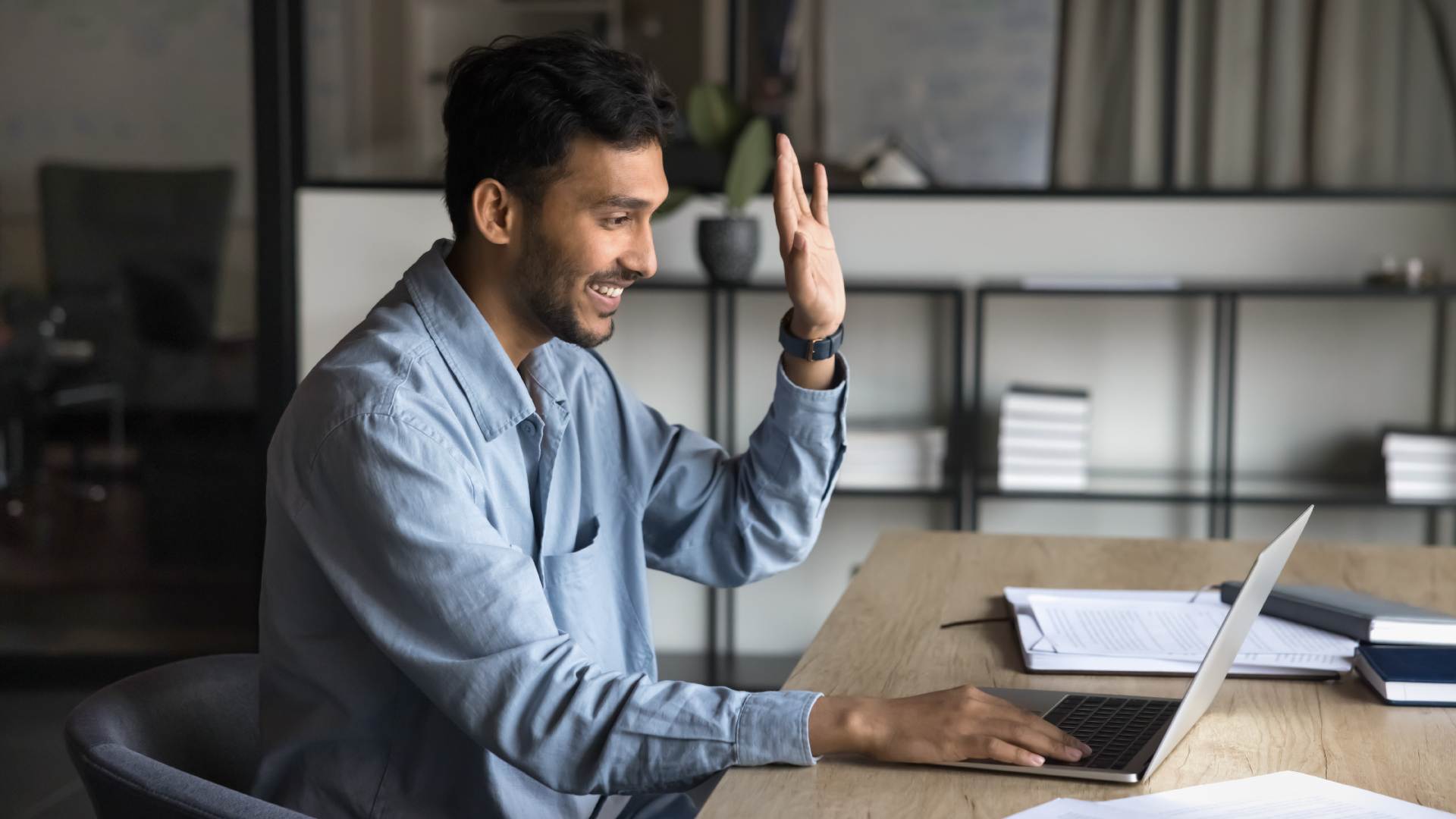 Smiling professional man waving during video call on laptop at home office desk