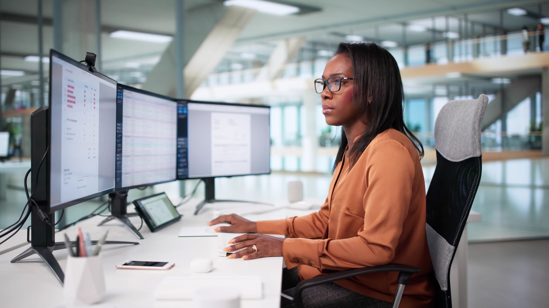 Professional woman with glasses working at triple-monitor desk setup in modern open-plan office